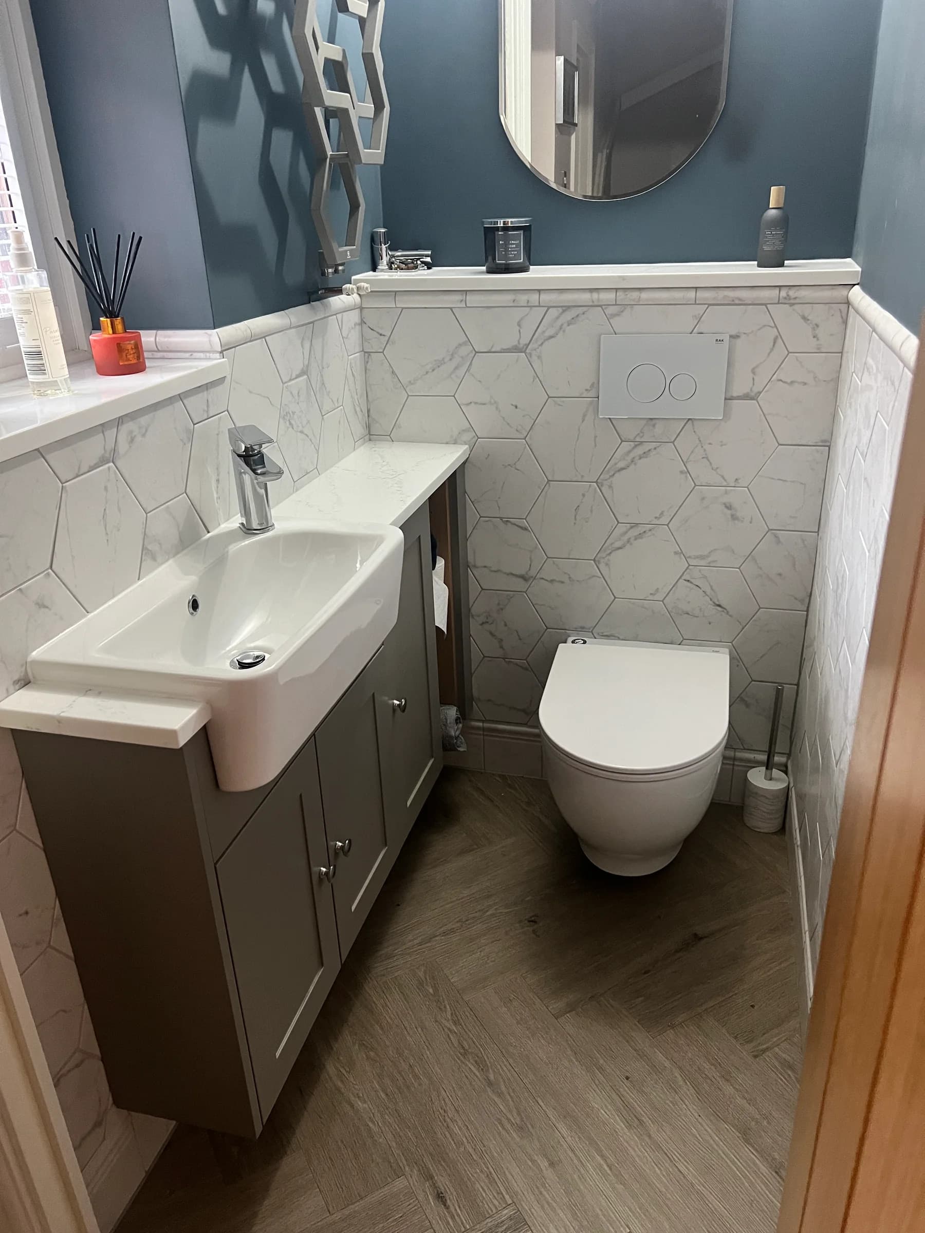 Compact bathroom installation with a basin cabinet, toilet, hexagonal wall tiles and a round mirror.