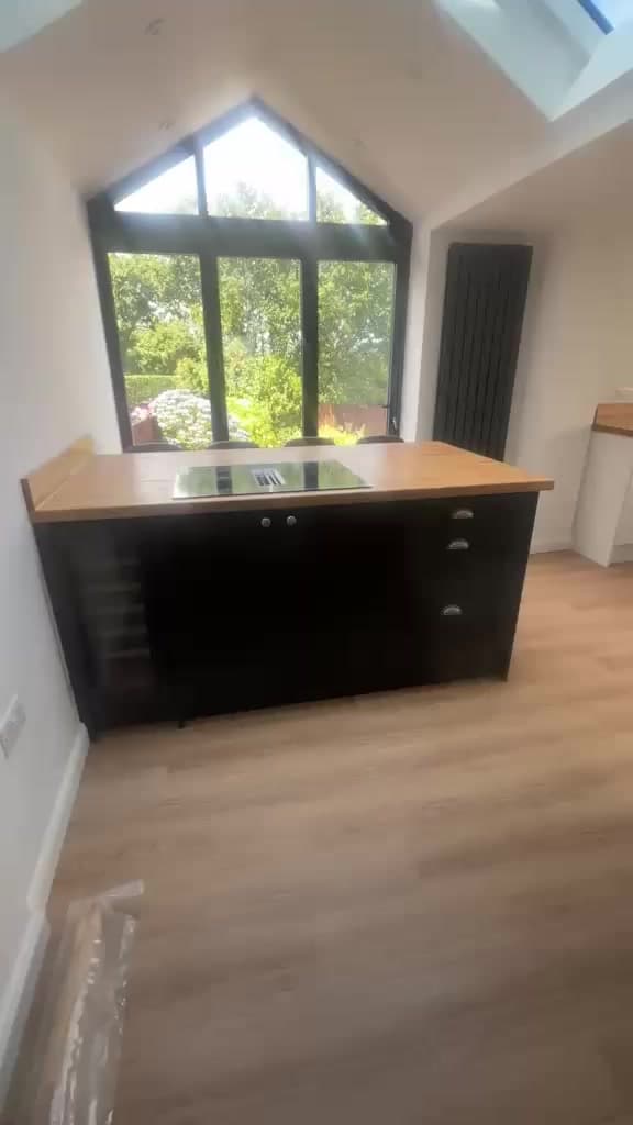 A kitchen island with dark cabinets and a wood worktop beneath a vaulted ceiling, with a garden visible through the glazing.